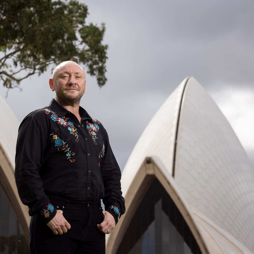 Beau James standing outside the Sydney Opera House. They're wearing a black shirt with floral details, and black pants.
