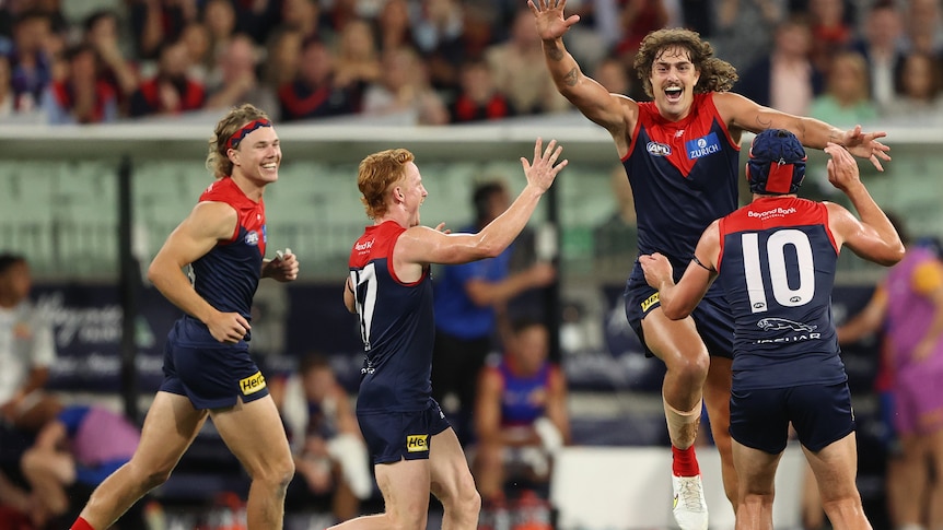 A tall AFL player grins as he leaps high in the air in celebration into the arms of his teammates.