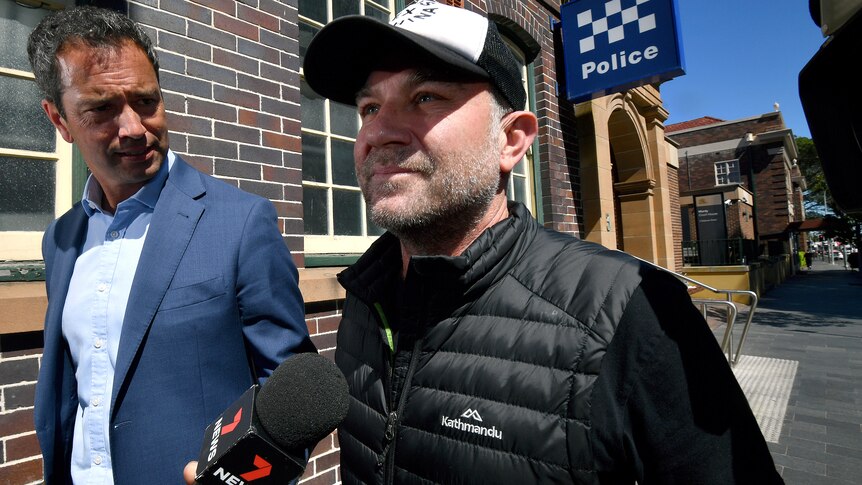A middle-aged man in a cap and puffer jacket walks next to a journalist outside a police station.