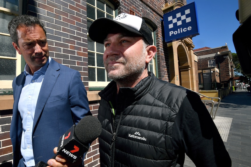 A middle-aged man in a cap and puffer jacket walks next to a journalist outside a police station.