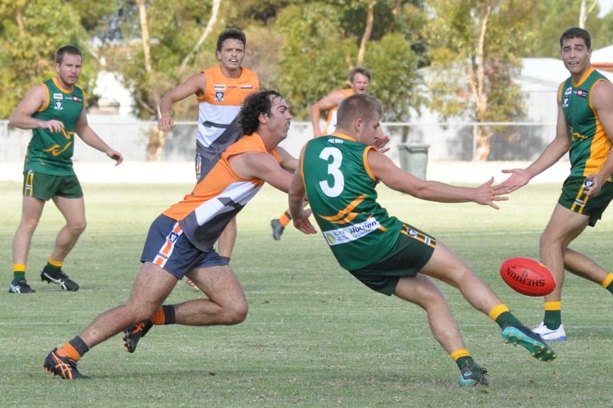 A footballer in a green jersey kicks the ball while a footballer in an orange jersey tries to smother the kick. 