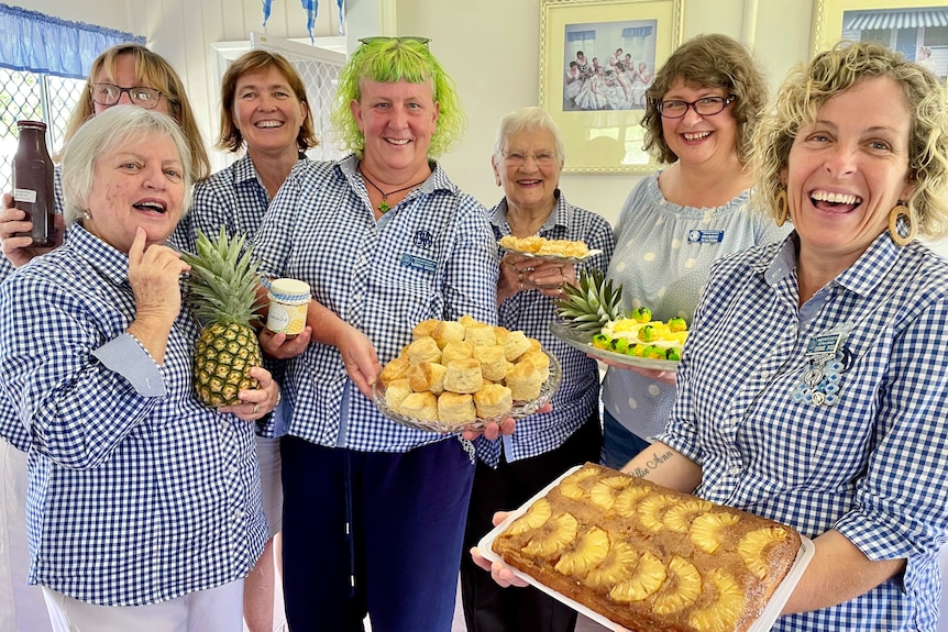 Women smile at the camera holding an array of tasty pineapple scones, cakes, jam and a fresh pineapple.