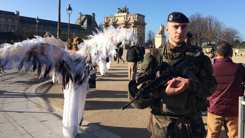 Climate change activists dressed as angels protest during climate talks in Paris, while a military officer stands nearby.