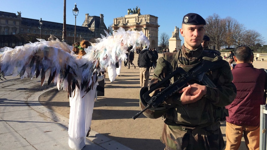 Climate change activists dressed as angels protest during climate talks in Paris, while a military officer stands nearby.