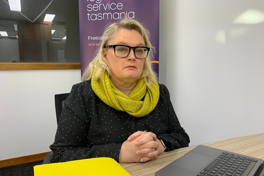 A woman looks seriously at the camera while sitting at a desk with yellow folders and a laptop.