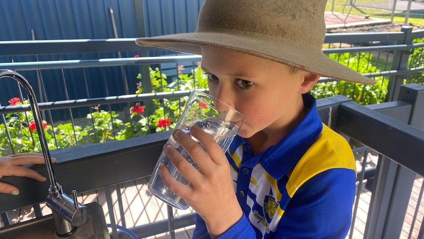 A primary school student in a blue uniform drinking water from a glass.