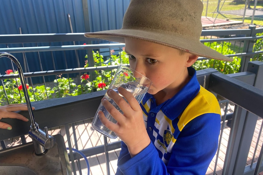 A primary school student in a blue uniform drinking water from a glass.