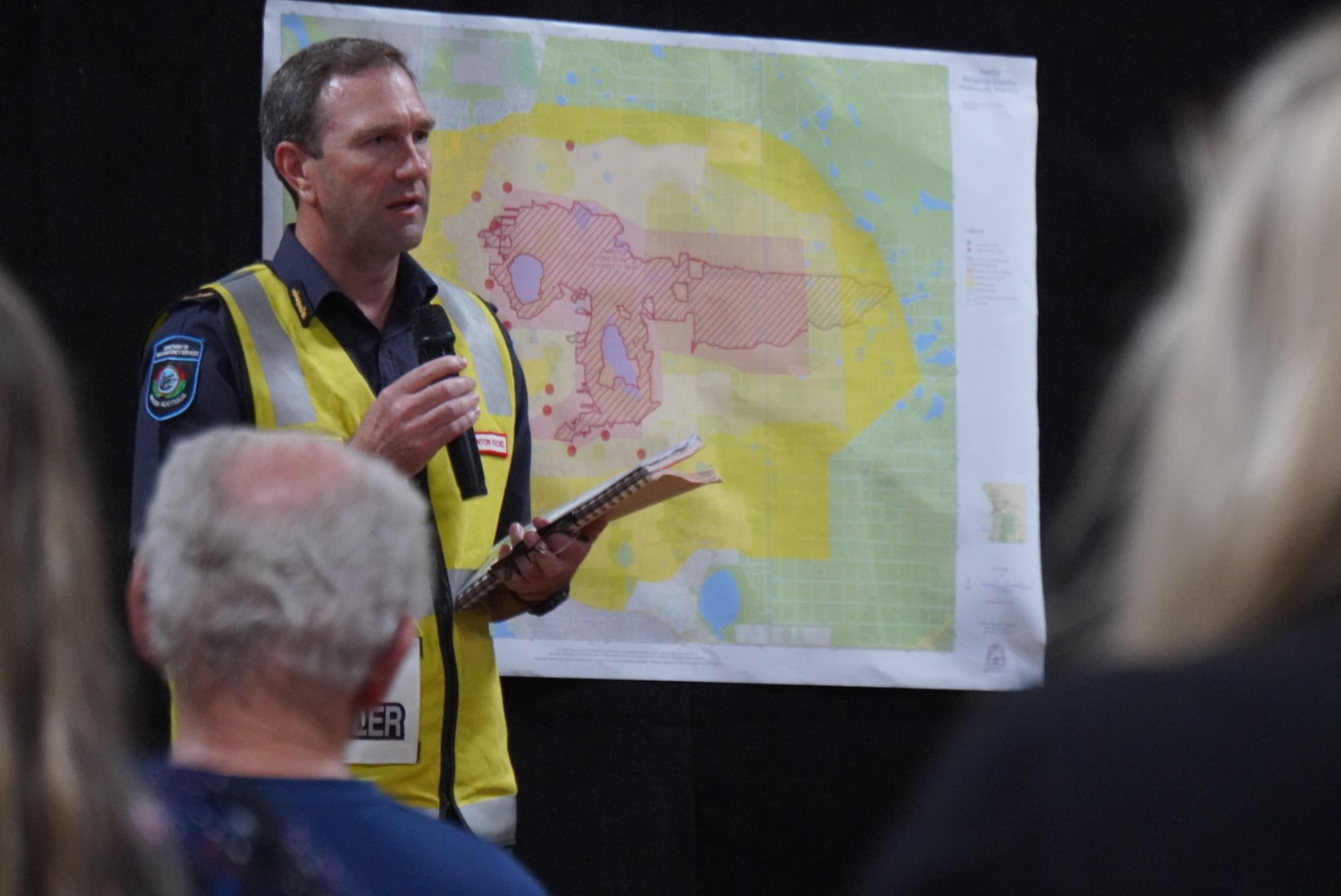 Um homem parado em frente a um mapa segurando um microfone e vestindo um colete amarelo enquanto se dirige aos presentes.