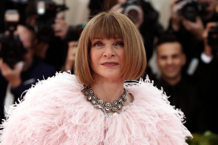 Anna Wintor smiles as she stands on the red carpet, photographers behind her. She wears a pink feather coat and diamond necklace
