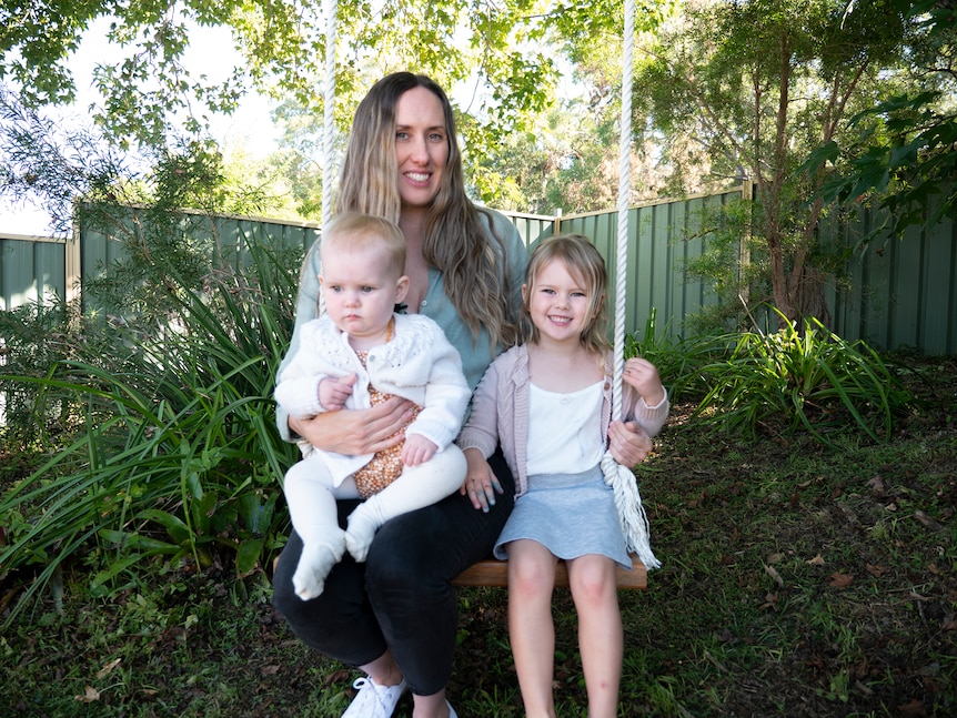 A woman and her two young female children sitting on a swing in a leafy environment.