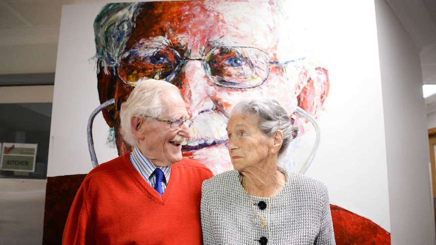 An elderly couple look into each other's eyes in front of the man's portrait.