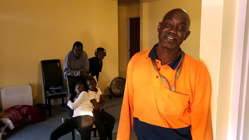 Arou Akot posing for a photo leaning against a wall of his unit while four sit and stand around a chair in the background