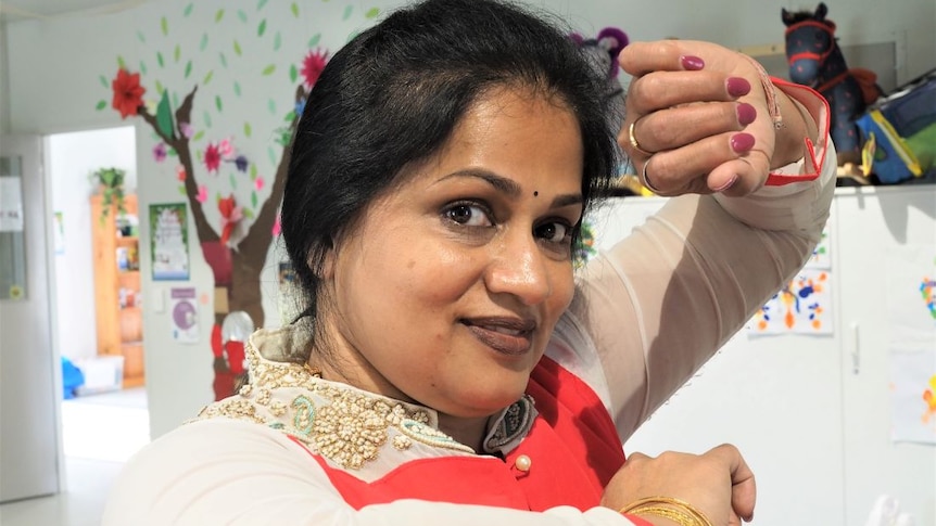 An Indian dance teacher poses with one arm in front of her forehead in a room filled with children's toys and art.