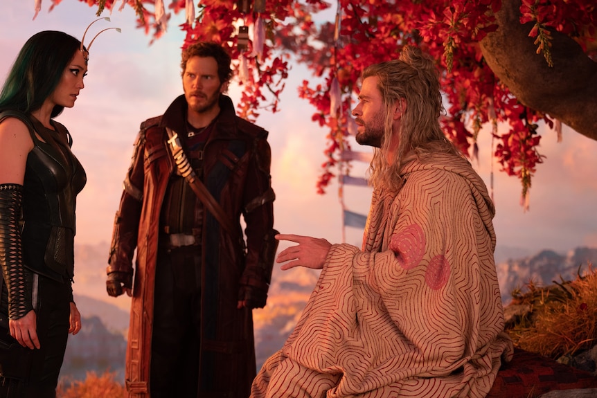 White man with long blonde hair wears white robes and sits outdoors under tree while talking to two others.