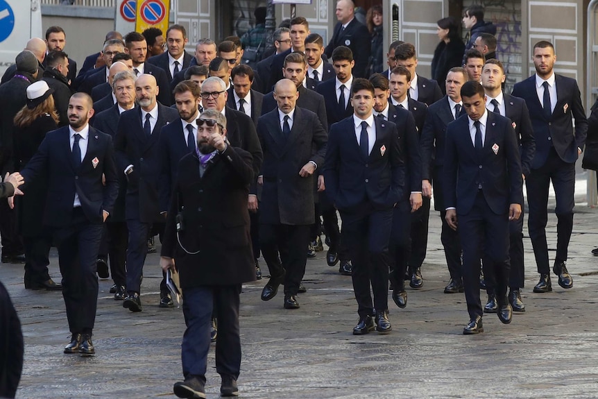A large group of Fiorentina players wearing black suits walk together.