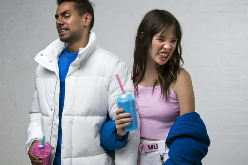 A young man and woman stand arm-in-arm with slushie drinks in their hands, grinning and baring rotten teeth.