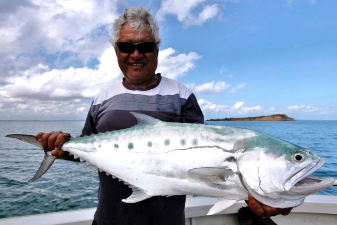 A close shot of Reuben Cooper standing in a boat holding a 1 metre long Queen fish. Blue water and sky surrounds.