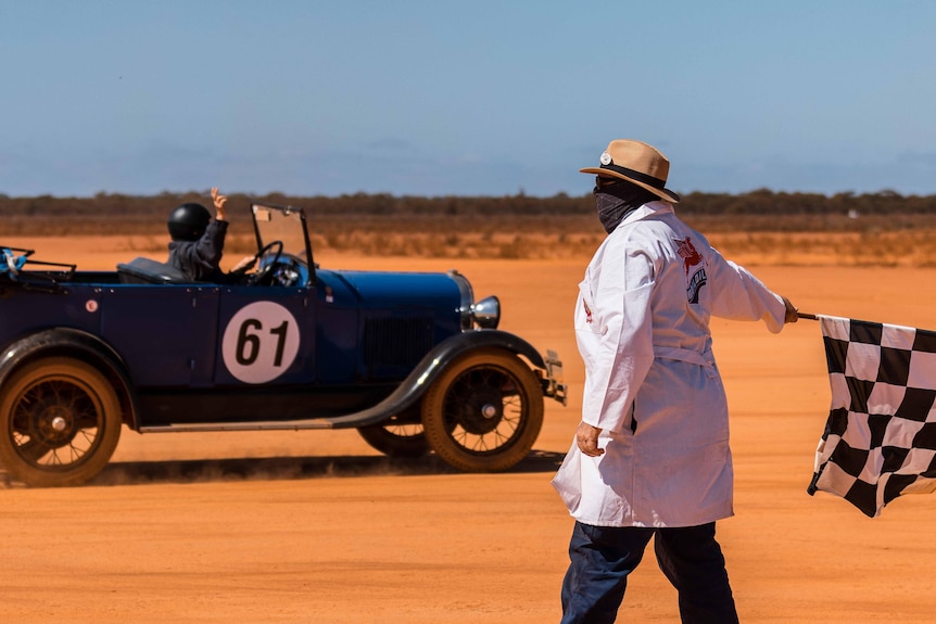 A man in a white coat waves the chequered flag at a vintage race car.  