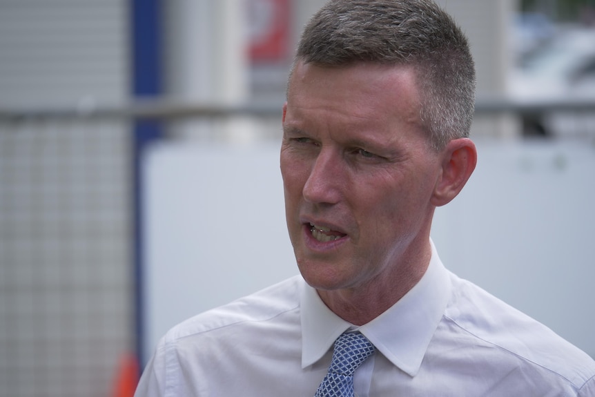 A middle-aged man in white shirt, blue tie, greying hair cropped close, speaks to someone off camera.