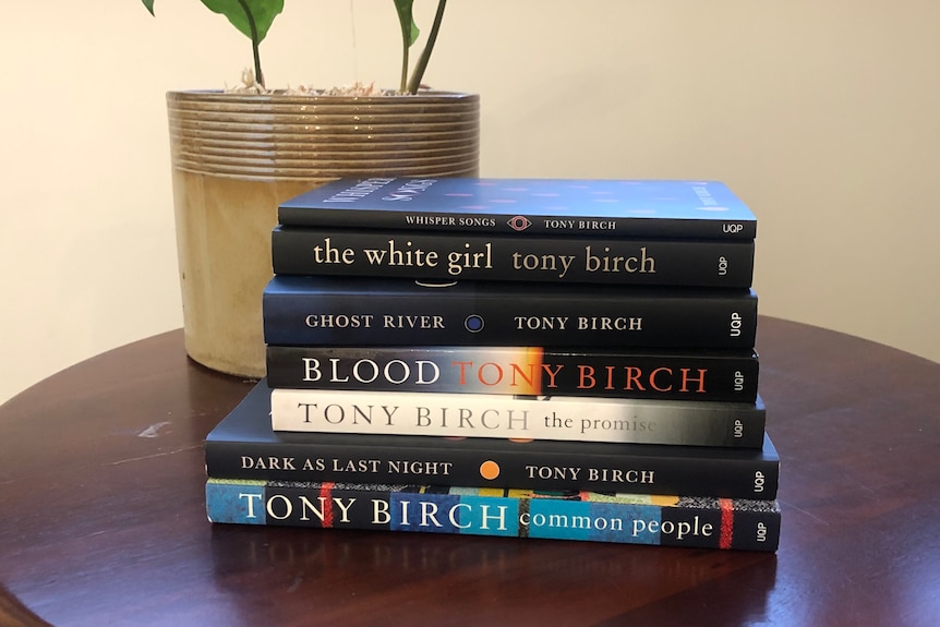 A stack of books by Tony Birch sits on a wooden table, a plant in the background