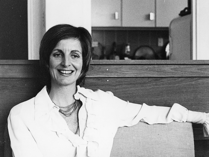 Caroline Jones smiles in a black and white photo, she's sitting, resting her arm on the top of the chair or couch.