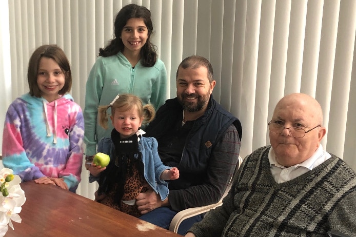 Three smiling children and a man with dark hair sit next to Apostolis Barbousas at a wooden table.