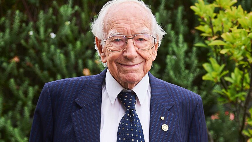 A mid-shot of a smiling 94-year-old David Bottomley posing for a photo in a blue suit and tie and white shirt.