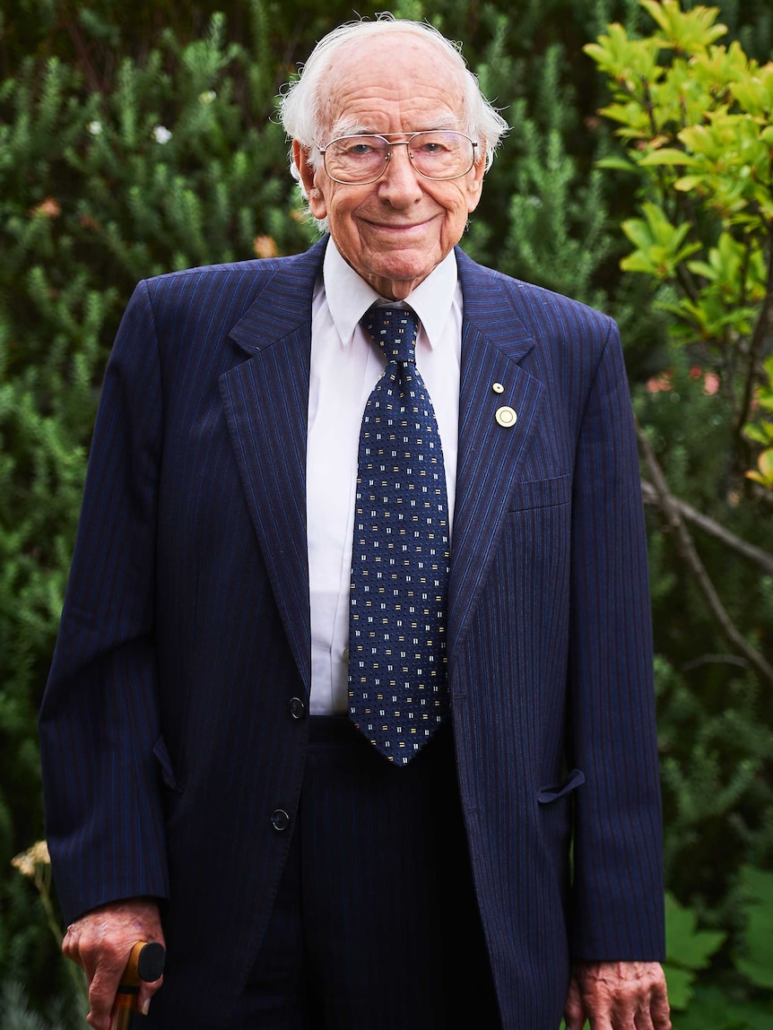 A mid-shot of a smiling 94-year-old David Bottomley posing for a photo in a blue suit and tie and white shirt.