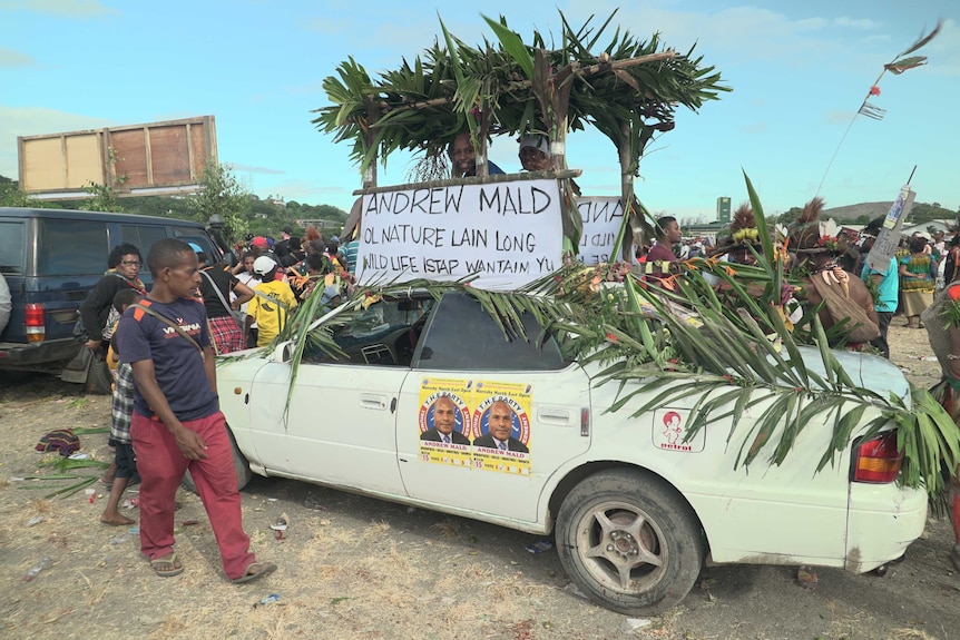 Two supporters smile under a thatched shelter sticking out of an Andrew Mald campaign car.