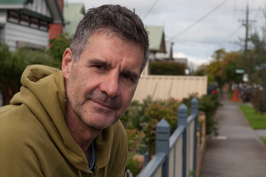 A man in a hooded jumper leans over a front yard fence