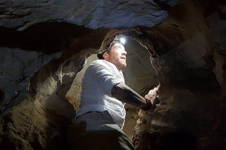 A man in a white shirt with a head torch is looking up inside a small cave.