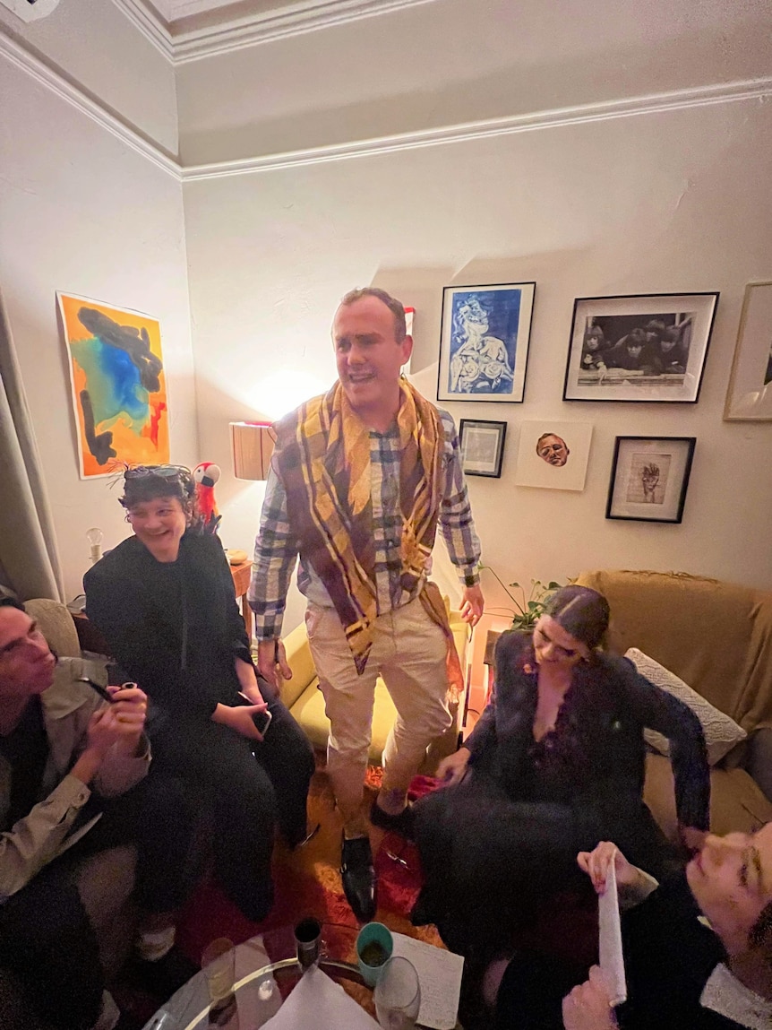 A man stands in a loungeroom surrounded by friends.