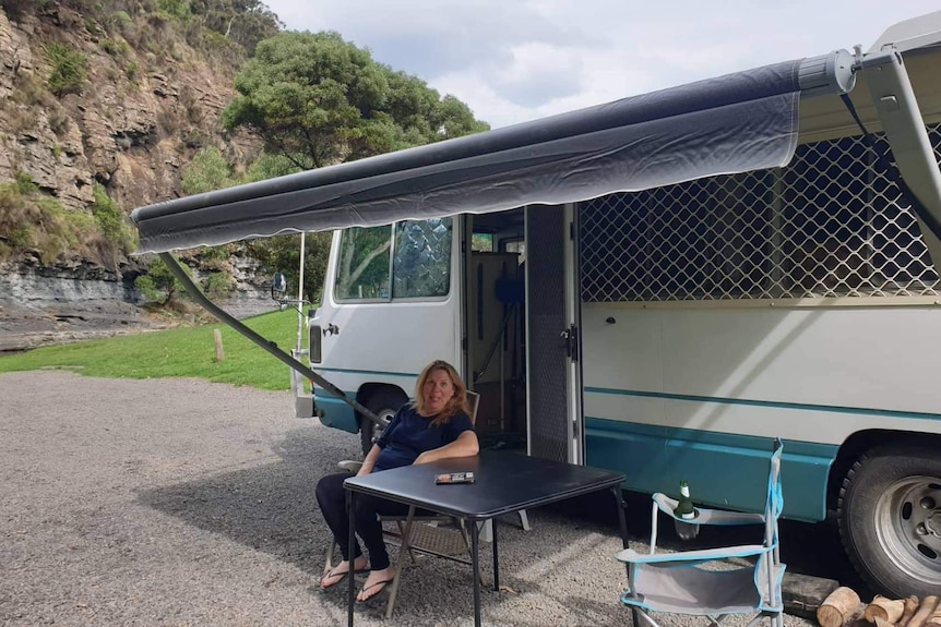 A woman sits in front of a bus with cliffs behind her.