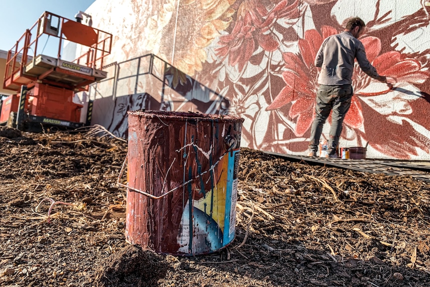 a close up image of an empty paint bucket with an artist and mural in the background