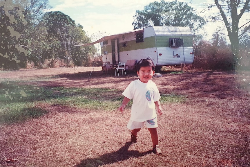 A toddler is seen walking towards the camera and smiling in front of a caravan at the edge of a farm. The grass is brown.