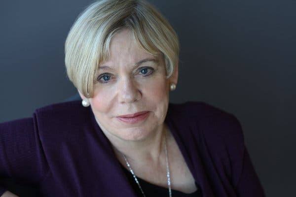 A close up photo of Karen Armstrong, who has short blonde hair, blue eyes and is wearing pearl earrings