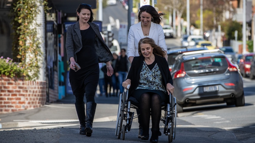 A woman in a wheelchair accompanied by two younger women on a sidewalk.