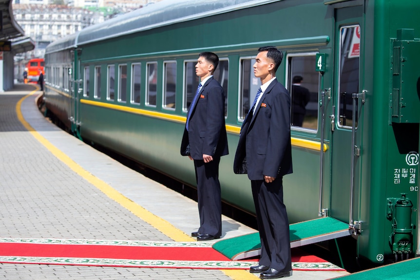 Two security guards stand to attention outside the door of a green train at a train station.