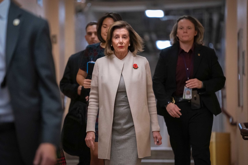 Nancy Pelosi walks down a hall way with several people walking behind her