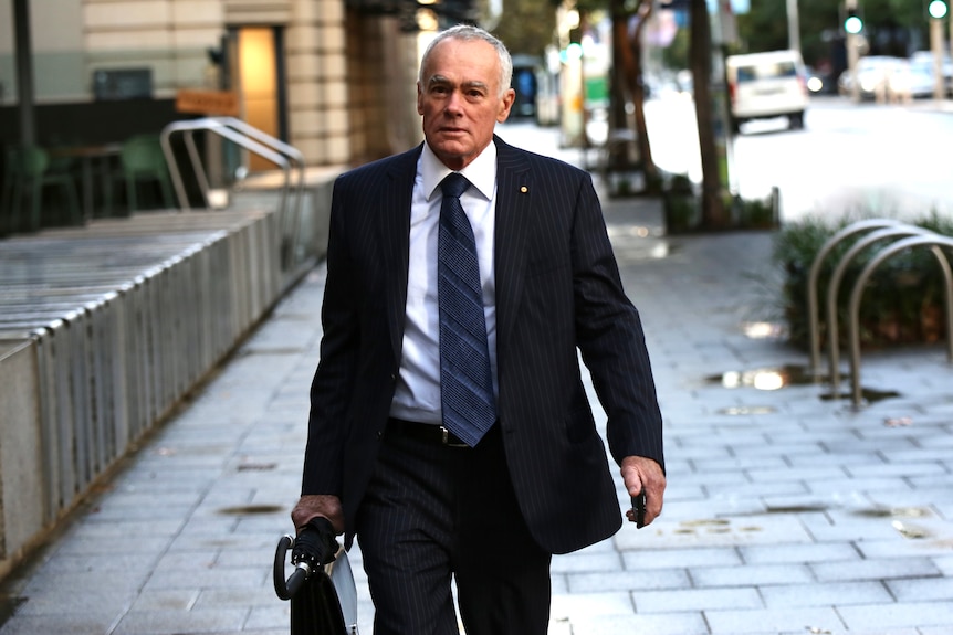 A mid-shot of John Poynton walking outdoors in the Perth CBD wearing a suit and tie.