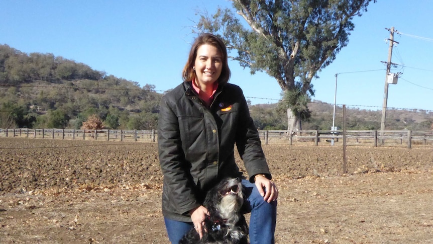 A woman with brown hair standing in a dirt paddock with a black dog.