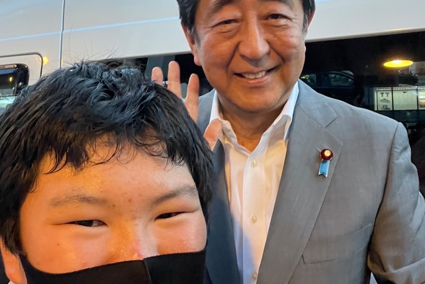A young boy wearing a mask poses for a selfie with a smiling Shinzo Abe