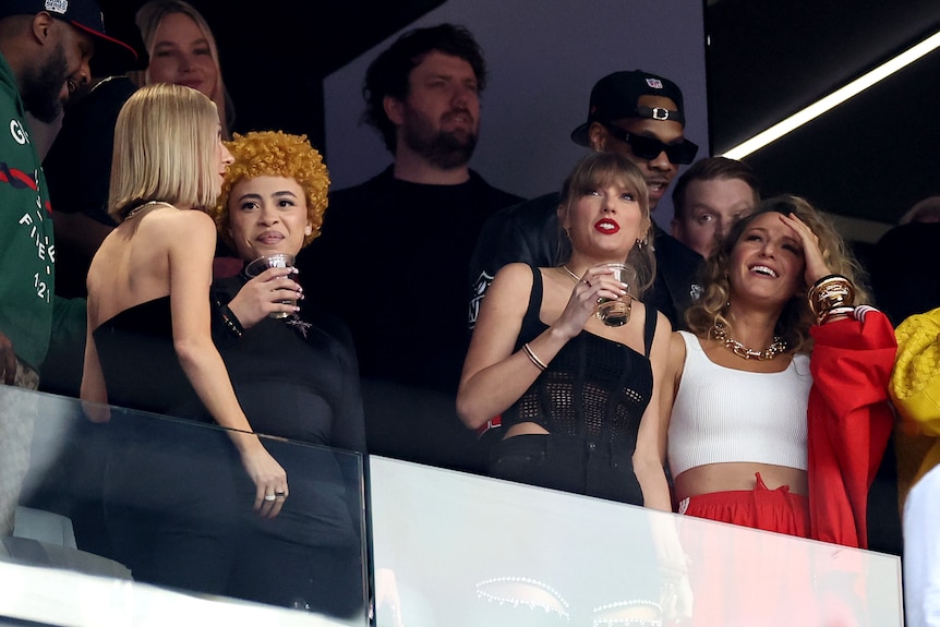 Taylor Swift wearing black, with Ice Spice next to her and Blake Lively in red and white on the other side, standing in stands
