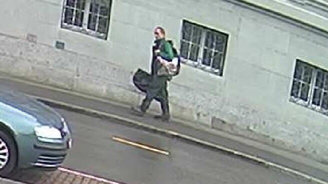 Grainy CCTV footage shows a man carrying two large bags walking down a street.