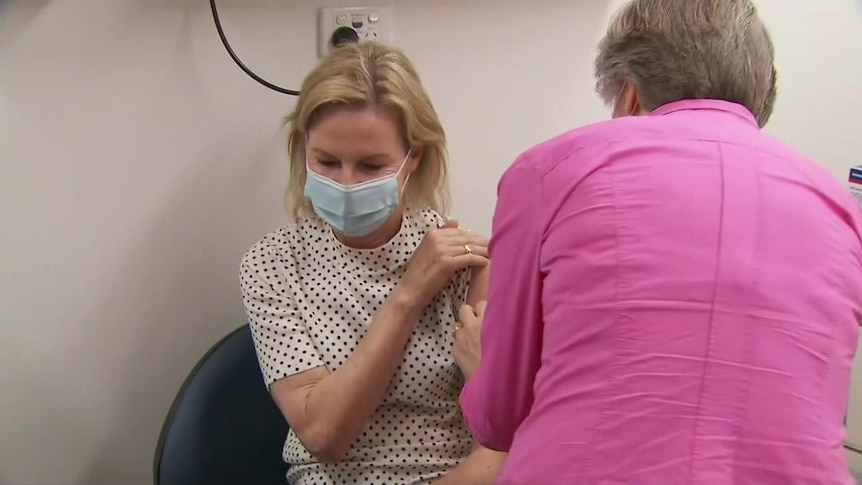 a blonde woman being vaccinated by another fair-haired woman in a pink shirt with her back to the camera
