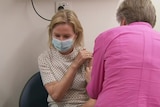 a blonde woman being vaccinated by another fair-haired woman in a pink shirt with her back to the camera