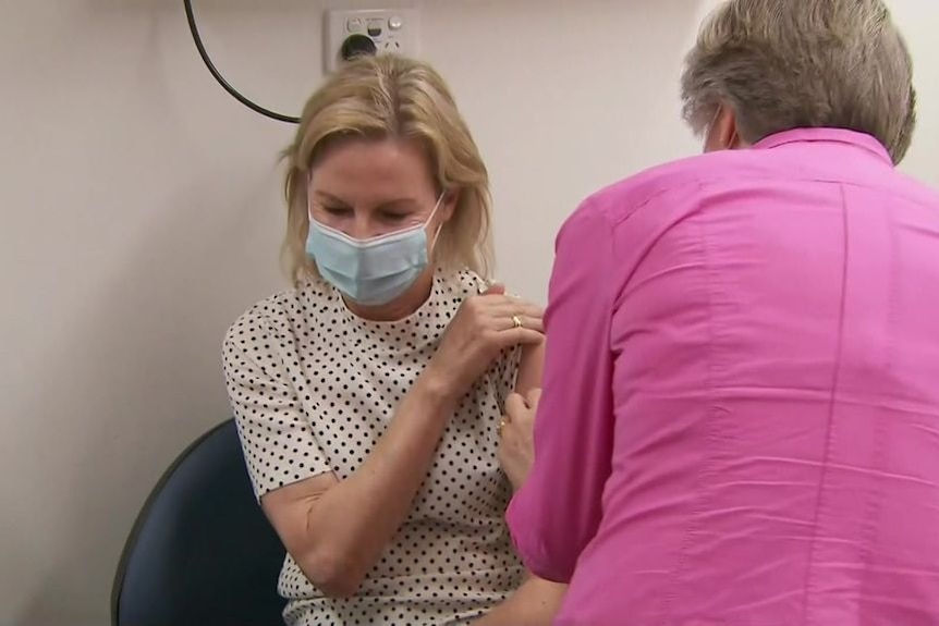 a blonde woman being vaccinated by another fair-haired woman in a pink shirt with her back to the camera