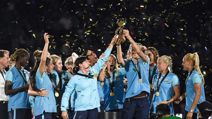 Australia's women's soccer team lifts the Tournament of Nations trophy as confetti falls on them.