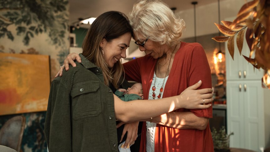Two women, one with grey hair, one with brown hair, in a kicthen, hugging with baby between them