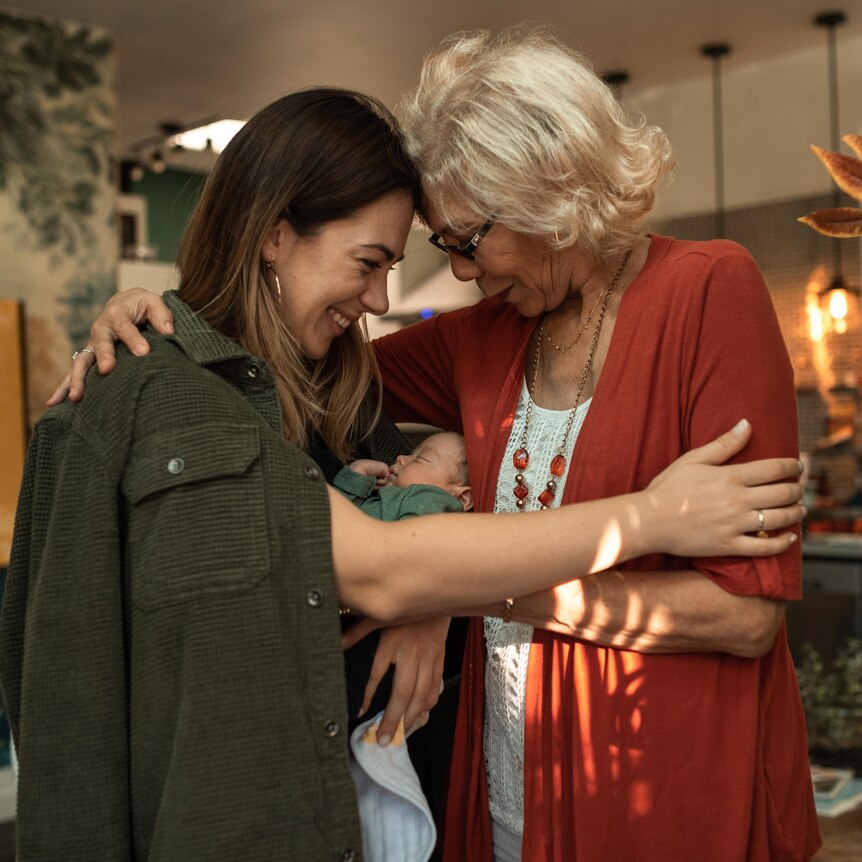 Two women, one with grey hair, one with brown hair, in a kicthen, hugging with baby between them